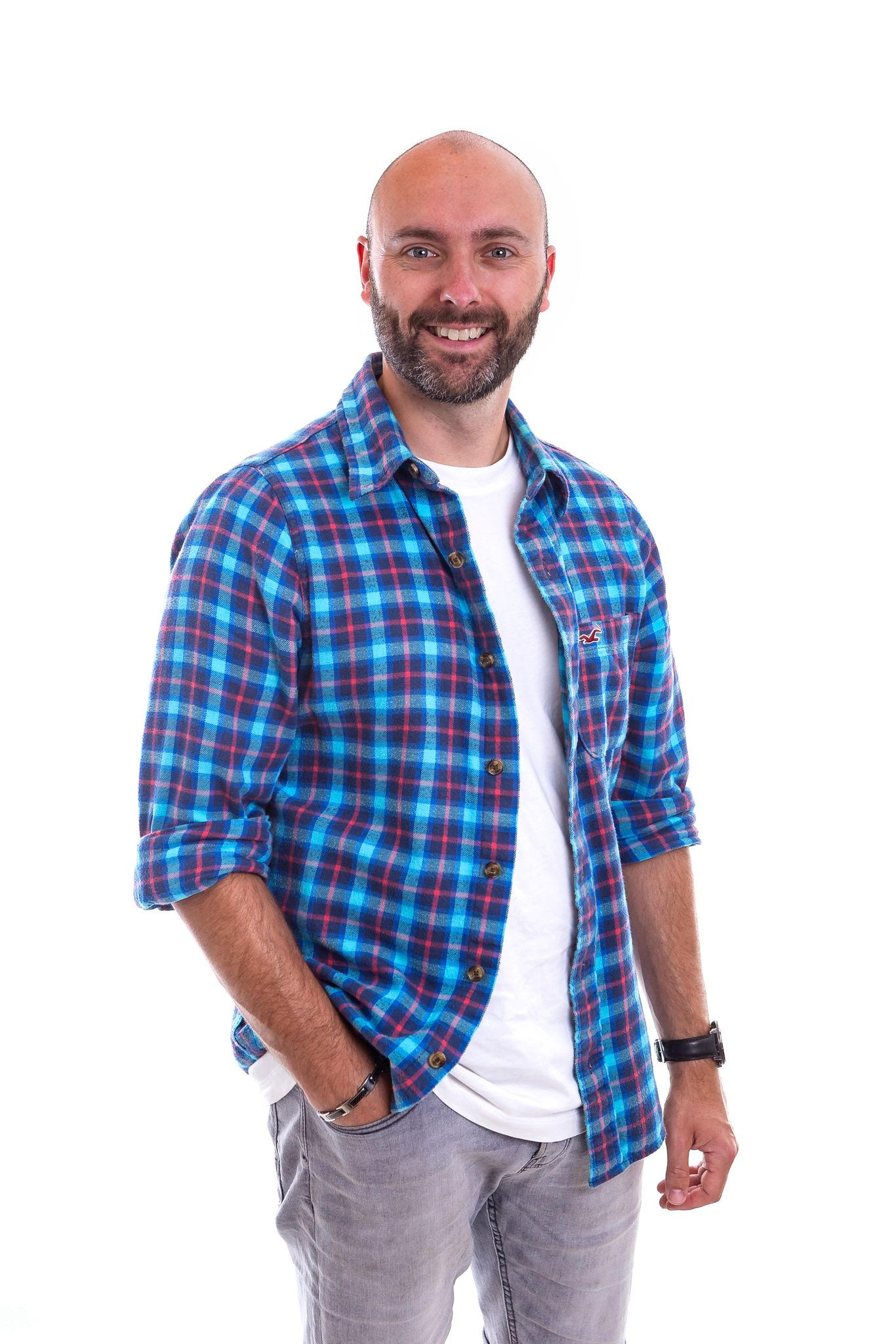 A smiling man with a beard wearing a blue and red plaid shirt over a white t-shirt, standing against a plain white background.